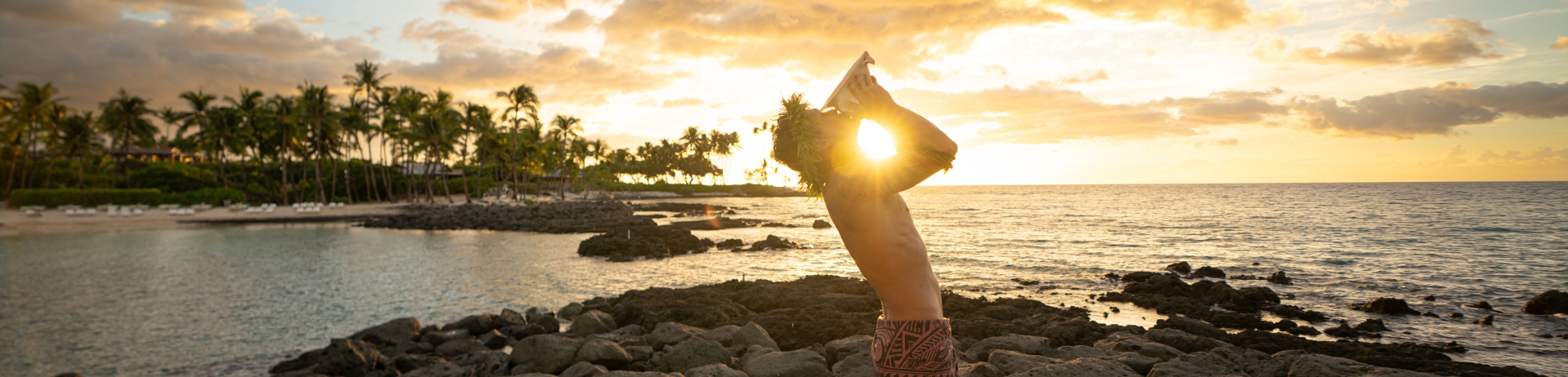 Man standing by the ocean blowing a conch shell at sunset